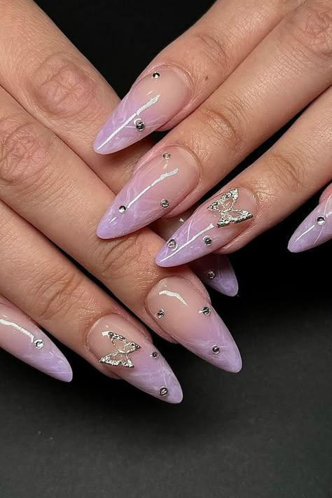 Elegance meets sparkle with these stiletto-shaped purple nails. The sheer lavender base exudes a soft, feminine aura, complemented by graceful marble accents that add depth. Each nail is embellished with tiny rhinestones, leading to larger, intricately designed silver butterflies that appear to gently alight on the canvas. This design embodies a subtle yet striking beauty that's sure to charm.  // Photo Credit: Instagram @lolliesbeautiq Purple Nails Crystals, Cute Feminine Nails, Purple Nails Elegant, Purple Birthday Nails Almond, Acrylic Butterfly Nail Designs, Purple Nail Designs With Butterflies, Almond Purple Nails Designs, Gel Nails Ideas Butterfly, Buterfluffy Nails Design