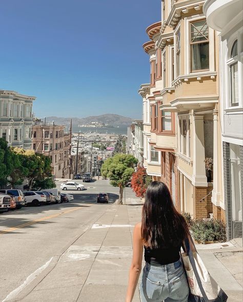 San Francisco painted ladies, antique buildings along the street facing a slanted hill. There’s a view of the city scape and a girl walking down the hill with the picture taken from her back. She has long black hair, jeans, and a trader joes tote bag. Sanfransisco Street Aesthetic, Cute Outfits For San Francisco, San Francisco Spring Break, Traveling To San Francisco, Sf Instagram Pictures, San Francisco Astethic, San Francisco Aesthetic Photos, Apartment In San Francisco, Life In San Francisco