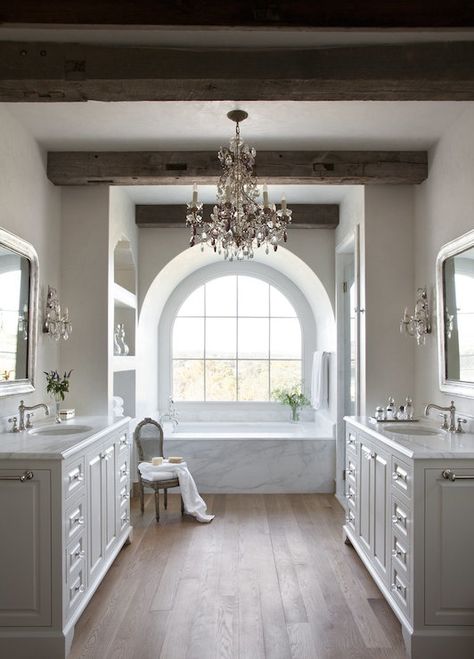 Stunning master bathroom centers on an arched alcove filled with arched window over a large soaking tub framed by a white marble surround flanked by built-in shelving to the left and a glass walk-in shower to the right below rustic wood beam ceilings adorned with a large crystal chandelier. Master Bathrooms, Rustic Glam, Dream Bathrooms, Dream Bathroom, Transitional Decor, Bathroom Remodel Master, Beautiful Bathrooms, White Bathroom, My New Room