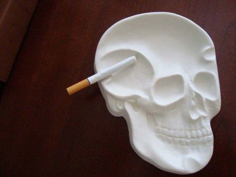 a cigarette sits along the side of a white ashtray. The ashtray is made out of clay, and is shaped like a 2-dimensional human skull. Air Dry Clay Ashtray, Skull Ashtray, Diy Skeleton, Kitchen Counter Top, Skull Wedding Ring, Skull Engagement Ring, Skull Pin, Skeleton Skull, Ceramic Bottle