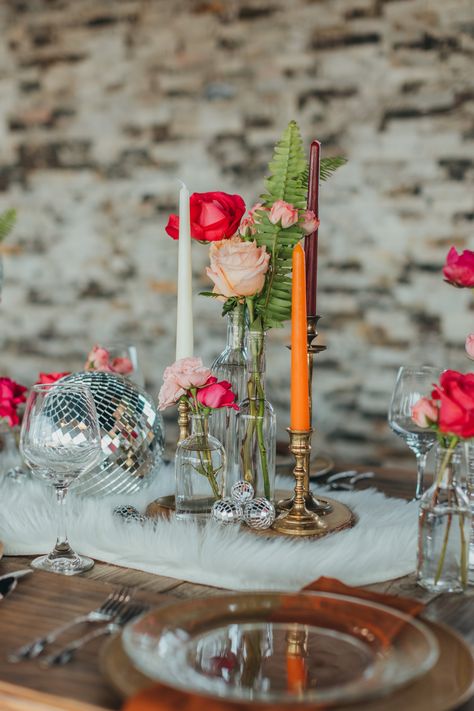 We had so much fun styling this boho disco themed shoot! We used vintage elements in combination with classic boho decor to make a super fun and unique table. These long farm tables are great for styling unique centerpieces. For ours, we used a combination of vintage candlestick holders, bud vases filled with assorted pink flowers, shiny disco balls, and went with a fun fur throw as a table runner. Sparkly and metallic gold chargers looked just right with a warm brown napkin. Shot by @kaboovang Disco Ball Wedding Tables, Fun Flower Vases, Centerpieces With Disco Balls, Pink Bud Vases Wedding, Disco Centerpieces Wedding, Disco Wedding Flowers, Disco Ball Wedding Table Decor, Disco Wedding Table Setting, Fall Disco Wedding