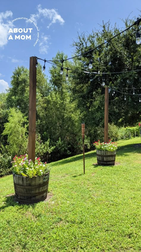 DIY Planter with Pole for String Lights - About a Mom Wood Poles For Outdoor String Lights, Front Yard String Lights, Acreage Yard Ideas, Wine Barrel Planter With Lights, Concrete Bucket String Lights, Planters With Poles For Lights, Diy Light Pole Planter, Pole For String Lights, Wine Barrel Lighting