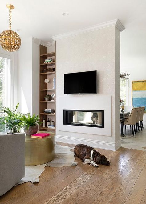 Living room designed with a double sided fireplace with white agate pattern wallpaper displaying a wall mount TV flanked by a built-in wood shelf. Double Sided Tv Wall, Double Sided Fireplace Living Room, Living Room Zones, Separation Wall, Mount Tv, Two Sided Fireplace, White Marble Bathrooms, Coastal Cottage Style, Rooms Design