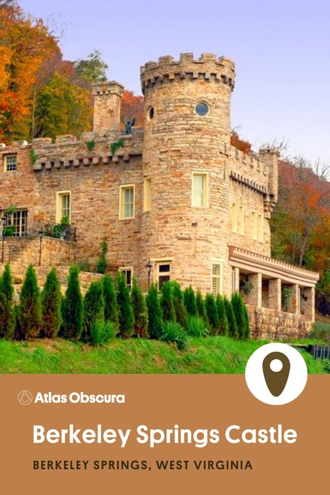 A sand-colored castle on the side of the road with a tower, chimney and patio surrounded by shrubs, trees and green grass. Fall leaves are in the background. Berkeley Springs Castle in Berkeley Springs, West Virginia.