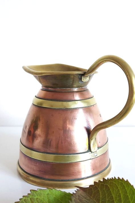 A copper jug with brass rings and spout is standing on a white surface, with leaves to the foreground. The jug is at a diagonal angle with the spout facing left and the handle right. Dried Flower Vase, Brass Pitcher, Vintage Jugs, Brass Rings, Vintage Pitchers, Copper And Brass, Brass Ring, Dried Flower, Flower Vase