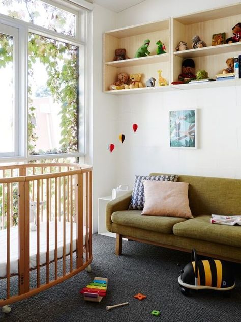 a beautiful mid century modern nursery with open box shelves, a stained round crib, a green sofa, artworks, bright toys that double as decor Mid Century Modern Nursery, Mid Century Nursery, Round Cribs, Elegant Nursery, Modern Crib, North Melbourne, Modernist House, Modern Kids Room, Modern Nursery Decor