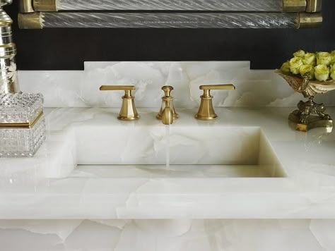 A white onyx vanity top is fitted with a brass faucet kit sat in front of an onyx backsplash and below a glass and brass vanity framed by black walls. Onyx Bathroom Ideas, Onyx Countertops, Custom Sinks, White Onyx, Vanity Countertop, Sink Countertop, Brass Faucet, Bathroom Countertops, Powder Bath