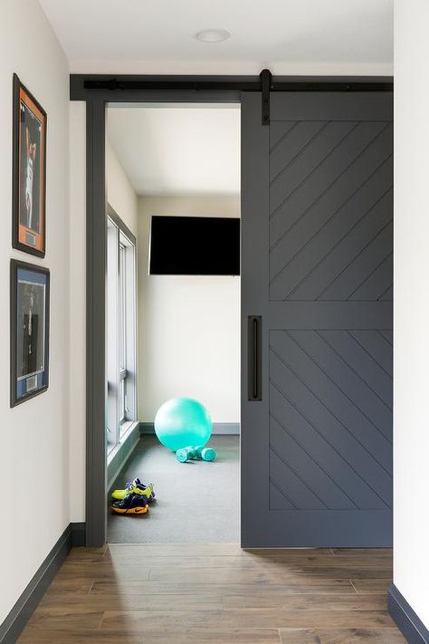 A dark gray plank barn door on rails is accented with a matte black handle and opens to a home gym boasting a flat panel television. Barn Door Basement, Basement Home Gym, Basement Gym Ideas, Rustic Basement, Basement Gym, Home Gym Decor, Black Barn, Home Gym Design, Sliding Doors Interior