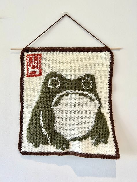 ✨Make your own Matsumoto Hoji frog crochet tapestry!✨ 📏 This tapestry is approximately 34cm x 36cm in size, measured from the bottom of the dowel rod 📏 ✍🏼This pattern includes instructions that have been written out in 3 different ways 1. Stitch Fiddle Graph  2. Colour Block Instructions  3. Written Instructions  🧶Materials needed:🧶 - 4mm crochet hook (can use other sizes at your own discretion) - Brown DK wool - Red DK wool - Dark green DK wool - Cream DK wool - Dowel rod - Scissors - Darn Plant Crochet Blanket, Crochet Hanging Shelf, Dog Tapestry Crochet, Frog Tapestry Crochet, Pokemon Tapestry Crochet, Crochet Pattern Tapestry, Halloween Crochet Tapestry, Small Crochet Tapestry Pattern, Crochet Tapestry Ideas