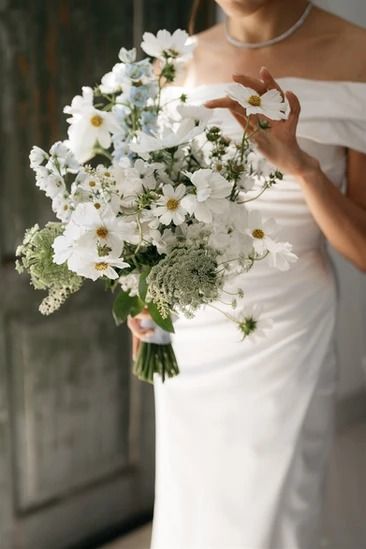 A bride stands indoors, smiling and looking down at her bouquet of white and light green flowers. She wears an off-shoulder white wedding dress and a delicate necklace. The background features a rustic wooden door and a green cabinet. Herb Flower Arrangements, White Bouquet Bride, White Wild Flowers Wedding, Airy Bridal Bouquet, Minimalistic Bouquet, White Garden Bouquet, White Wildflower Bouquet, Simple White Bouquet, Stock Bouquet