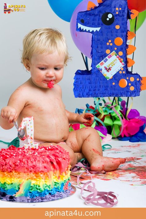 A baby celebrating their first birthday sits in front of a rainbow-colored cake, covered in frosting, while playing with a spoon. A blue dinosaur piñata with the number "1" and colorful balloons decorate the background. The website "apinata4u.com" is visible at the bottom. Dino Bday Party, First Birthday Blue, Number Pinata, Birthday Party Blue, Dino Theme, Dinosaur Theme, Let The Fun Begin, Dinosaur Party, Accessories Ideas