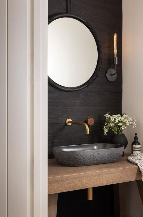 Small powder room nook features a black vessel sink on a wooden washstand under wall mounted brass faucet and a round black rope mirror on a black oak accent wall lit by a long sconce. Black Half Bathroom Modern, Powder Room Bowl Sink, Modern Industrial Powder Room, Minimalist Powder Room Design, Black Vessel Sink Bathroom Ideas, Powder Room Vessel Sink, Oak Accent Wall, Black Half Bathroom, Black Vessel Sink