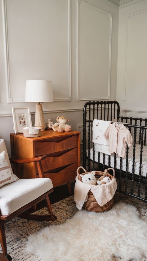 A sophisticated baby girl nursery with a black vintage-style crib, a mid-century modern wooden dresser topped with decor and a plush toy, a cozy rocking chair, a woven basket of stuffed animals, and a soft faux fur rug on a patterned floor. Baby Girl Nursery Black Crib, Black Crib Nursery Girl, Nursery With Daybed And Crib, Black Crib Nursery Ideas, Black Crib Nursery, Dusty Pink Nursery, Chic Baby Nursery, Vintage Baby Girl Nursery, Nursery Inspiration Girl