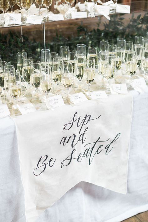 This custom "sip and be seated" matched the adorable placards attached to each glass so as they were seated they could already enjoy themselves. Accented with an acrylic pillar stuffed with greens and it gave us some added height and dimension Sip And Seat Wedding, Take A Sip And Take Your Seat Wedding, Grab A Drink And Take A Seat Sign, Sip And Sit Wedding, Sip And Be Seated Wedding, Welcome Drink Wedding, Champagne Seating Chart, Champagne Seating, Sip And Be Seated