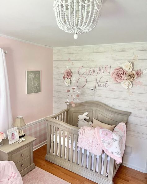 Pink and white flowers hang on a rustic white shiplap baby girl nursery wall above a light grey crib. A grey picture hangs on a pink wall with pink and white checkered wainscoting. A light grey end table is placed on a pink rug on the golden wood floor. Grey Crib Nursery, Rustic Girl Nursery, Shiplap Nursery, Shiplap Wallpaper, Gray Nursery Girl, Pink Baby Nursery