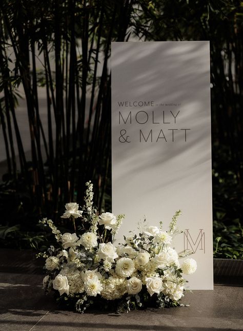 White modern wedding welcome sign with white flower arrangement at the base Florals For Wedding Sign, Welcome Sign Wedding With Flowers, Welcome Sign Flower Arrangement, Wedding Signing Table Flowers, Wedding Welcome Sign Floral Arrangement, Welcome Sign For Wedding Reception, Elegant Welcome Sign, Classic Wedding Welcome Sign, Wedding Seat Sign