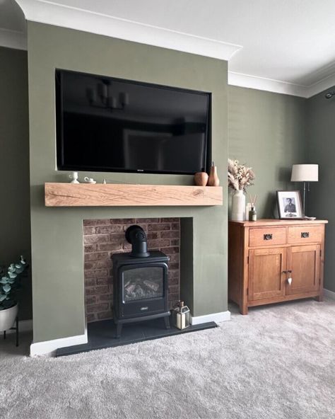 A living room with a green feature wall. The wall contains an alcove with an exposed stone interior housing a small black fireplace. Above the fireplace sits a solid wood mantel beam. To the right of the mantel beam are two vases, and to the left is a small china accessory. Mounted above the mantel beam is a built-in television. Adjacent to the fireplace is a wooden cabinet adorned with a picture frame, a lamp, a diffuser, and a vase filled with feather stems. Plank Fireplace Wall, Freestanding Fireplace Ideas, Wood Burning Fireplace Ideas, Warm And Cozy Aesthetic, Green Living Room Inspiration, Chunky Furniture, Sage Living Room, Tv Above Fireplace, Calm Green