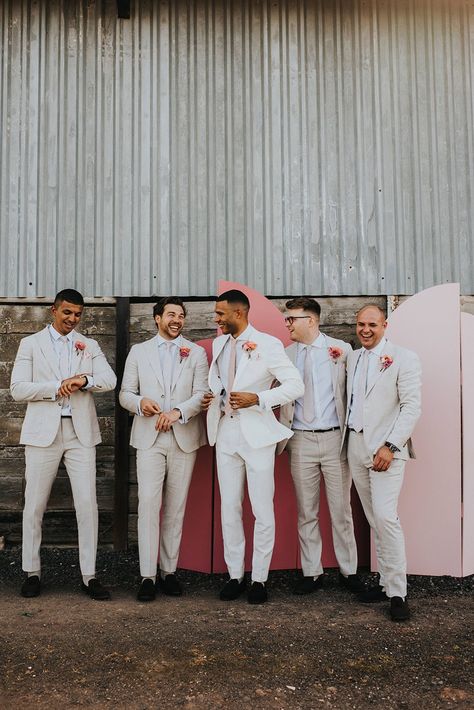 Groom in a white suit and groomsmen in cream suits with pink ties and pink flower buttonholes in front of the pink wedding screen background Light Grooms Suit, Bright Wedding Groomsmen, White And Pink Mens Suit, Blush Pink Groomsmen Suit, White Suit With Pink Tie, Groom Blush Wedding, Pink Grooms Suit, Pink Wedding Bride And Groom, Tan And Pink Bridal Party