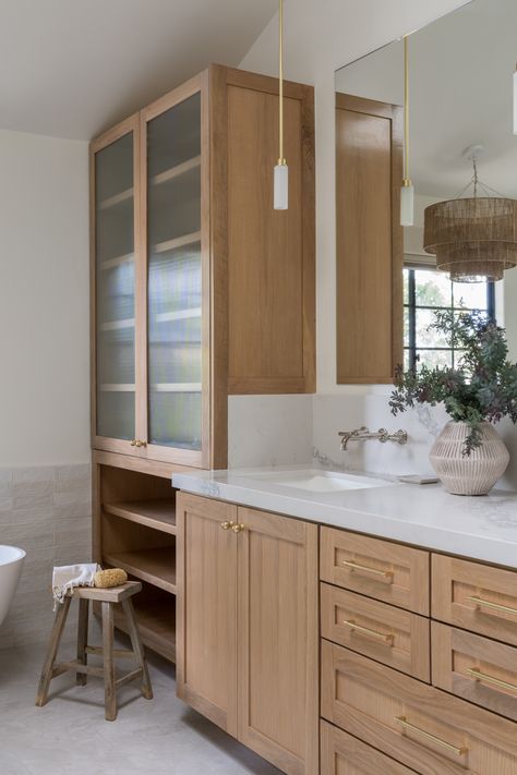 This primary bathroom transformation has all the textural elements that make it a spa-like retreat! White oak vanity, marble-look quartz countertops, limestone flooring, brass hardware, and reeded glass detail in the linen closet. White Oak Cabinets Brass Hardware, Glass Front Linen Cabinet, White Oak Bathroom Vanity Brass Hardware, Reeded Glass Bathroom Cabinet, Reeded Oak Vanity, Fluted Glass Bathroom Cabinet, Glass Upper Kitchen Cabinets, Calcutta Marble Bathroom, Glass Bathroom Cabinet