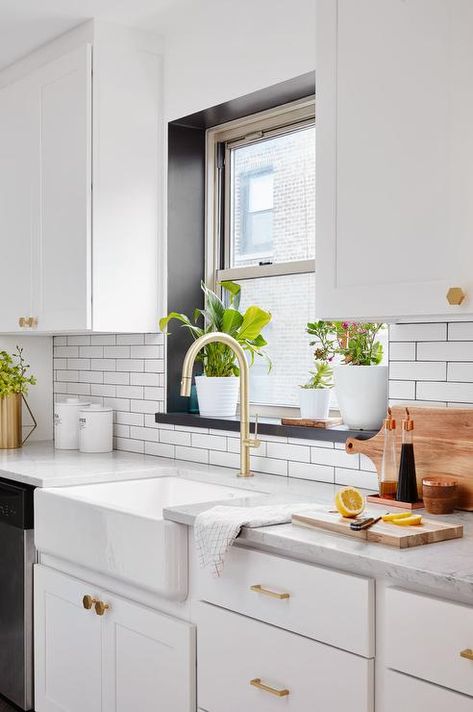 White and gold kitchen features a window framed by a black windowsill positioned above a farmhouse sink with a brushed brass gooseneck faucet fixed to a marble countertop placed against a white subway tile backsplash and on top of white shaker cabinets donning brass hexagon knobs. Window Over Sink, Farmhouse Sink Faucet, Kitchen Window Sill, Farmhouse Backsplash, Easy Home Improvement, White Subway Tile Backsplash, Herringbone Backsplash, Window Seal, Galley Kitchen