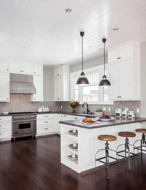 Contemporary kitchen features a beadboard ceiling over a white shaker cabinets adorned with oil rubbed bronze hardware and a gray quartz countertops, Caesarstone Lagos Azul,  and a gray mosaic tile backsplash. A pair of industrial pendants hang over a kitchen peninsula fitted with end shelves lined with backless wood and iron counter stools. Peninsula Kitchen, Kitchen Peninsula, Herringbone Backsplash, Condo Ideas, Garage Conversion, White Kitchen Design, Grey Kitchen, Trendy Kitchen, Kitchen Redo