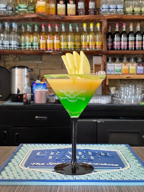 A martini glass with a skull on it is pictured. The liquid inside is layered- vibrant green on the bottom, caramel colored brown on top. The drink has a green apple garnish. Caramel Apple Sucker Drink, Green Apple And Caramel Vodka, Green Apple Vodka Drinks Recipes, Green Halloween Cocktail, Green Apple Caramel, Sour Green Apple Cocktail, Caramel Apple Suckers, Caramel Apples Halloween, Apple Syrup