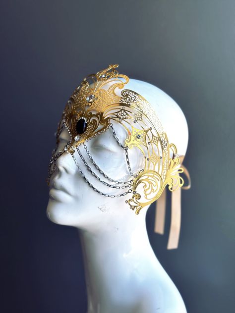 Step into the realm of goddesses with our women's masquerade mask in gold. Rhinestones delicately embellish the mask, adding a touch of sparkle. Silver chains gracefully adorn the mask, creating an enchanting movement. Capture attention at any masquerade party, ball, or themed event!   Age Group/Gender - Adult/Women  Size/Type - One size fits all adults  Mask Color - Gold  Mask Material - Laser Cut Metal  Special Features - Chains and rhinestones Ball Room Mask, Fairy Masquerade Mask, Angel Mask Masquerade Ball, Warrior Mask, Fairy Mask, Vintage Mask, Gold Fantasy Masquerade Mask For Carnival, Masquerade Mask Aesthetic, Luxury Traditional Gold Masquerade Mask