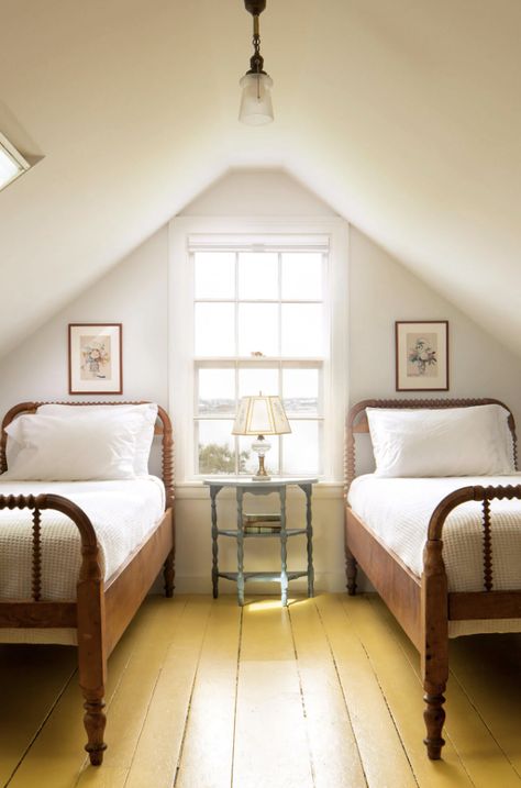 The attic space within this coastal Maine home was transformed into a cozy guest bedroom. Yellow painted floors set off the home's original twin spool beds. Framed art, a patterned lamp, and books compliment the painted bedside table, and create a calming aesthetic. White Attic Bedroom, White Attic, Tennessee Farmhouse, Vintage Kitchen Cabinets, Bedroom Boy, Southern Farmhouse, Antique Bed, Cottagecore Home, Slanted Ceiling