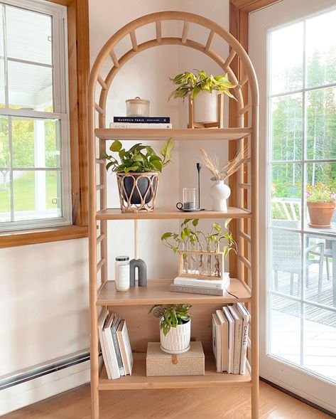 An arching rattan bookshelf is placed beside a bright paneled window, casting lots of natural light on the potted indoor plants. Wheatgrass, neutral books, and white candles accent the shelves along with the plants and wood vases for an ultra-neutral and organic look. Bamboo Room Decor, Arched Bookshelf Styling, Minimalist Bookshelf Styling, Bohemian Bookshelf, Boho Bookshelf, Boho Shelves, Bookshelf Decor Ideas, Rattan Bookshelf, Neutral Books