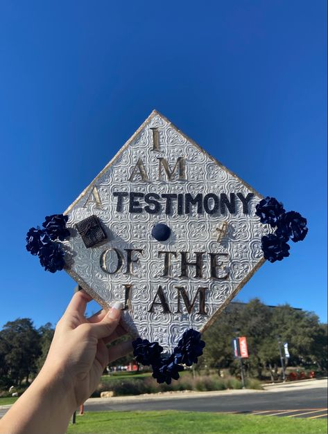 A decorated graduation cap reading “ I am a testimony of the I am.” Cap has cold trim with a white detailed background. Gold and black lettering. A small Bible on the left and cross on the right. Background is a college campus with a blue sky. Graduation Cap Christian Ideas, Graduation Cap Designs Jesus, Grad Cap Designs Christian, Godly Graduation Caps, My Story His Glory Grad Cap, Grad Cap Christian, Cap Decoration Graduation God, Cap Decoration Graduation Bible Verse, Catholic Graduation Cap