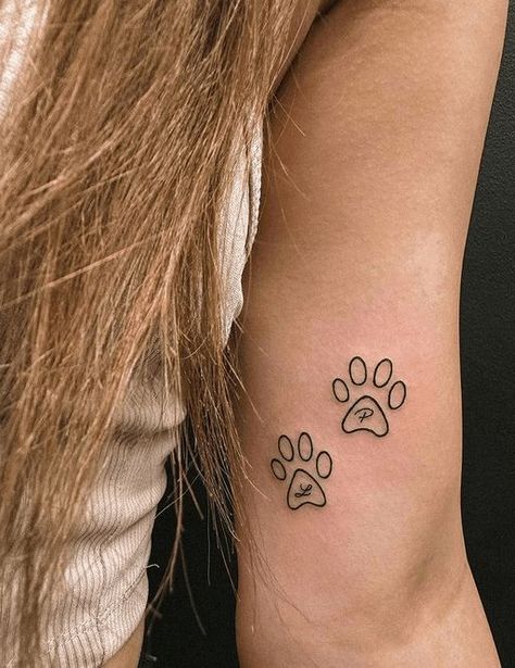 Animal Paw Print1 Two minimalistic paw print tattoos on a woman's upper arm, featuring initials L and P inside the prints. | Sky Rye Design Tattoos Paw Print, Print Tattoo Designs, Paw Print Tattoos, Watercolor Galaxy Tattoo, Animal Lover Tattoo, Cat Outline Tattoo, 40 Tattoo, Sky Tattoos, Basic Tattoos