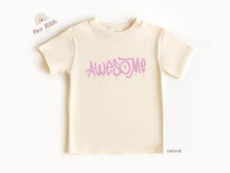 A cream-colored T-shirt with the word “Awesome” in bold, dripping pink letters, resembling a graffiti style. The font gives a playful and energetic vibe. Toddler Tshirt Ideas, Graffiti Rose, Cool Street Art, Pink Graffiti, Vibrant Personality, Graffiti Designs, Cool Graphic Tees, Graffiti Styles, Toddler Tees