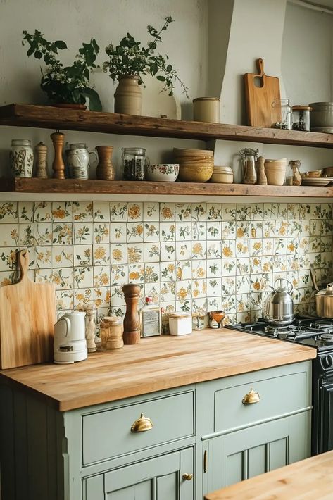 cozy cottage-style kitchen featuring a rustic wooden countertop and open shelving. The backsplash consists of floral-patterned tiles in soft yellow and green hues, complementing the sage-green cabinetry with brass hardware. The shelves are adorned with potted plants, ceramic jars, and vintage kitchenware, creating a warm and inviting atmosphere. Various kitchen essentials such as a kettle, pepper mill, and cutting boards add functionality to the charming design. Eclectic French Country Kitchen, Vintage Boho Decor Interior Design, Sage Green Kitchen Walls Cream Cabinets, Small Cottage Core Kitchen, Cottagecore Kitchen Cabinets, Cottage Kitchen Open Shelving, Cottagecore Backsplash, Cottagecore Kitchen Backsplash, Cottage Inspired Kitchen