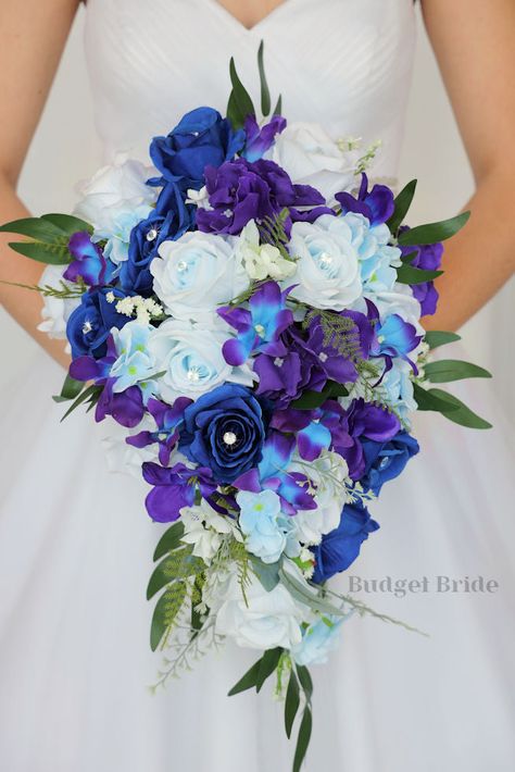 This lovely assortment of royal blue and light blue roses accented with galaxy orchids and luxurious greenery is bound to turn heads on your wedding day. This lovely cascading brides’ bouquet from the Moonstone collection is an absolutely breathtaking arrangement for any bride to be, in any season. Purple and blue hydrangea complete the look and add a delicate realism to this beautiful bouquet. This bouquet is 10” wide, 18” long, and is crafted by our in-house florists without using a foam base Blue Roses Wedding Decor, Royal Blue And Purple Bouquet, Royal Blue Purple And White Wedding, Blue Wisteria Bouquet, Royal Blue And Purple Wedding Dress, Royal Blue And Purple Wedding Ideas, Blue And Purple Bouquet Wedding, Royal Blue And Royal Purple Wedding, Galaxy Wedding Ideas