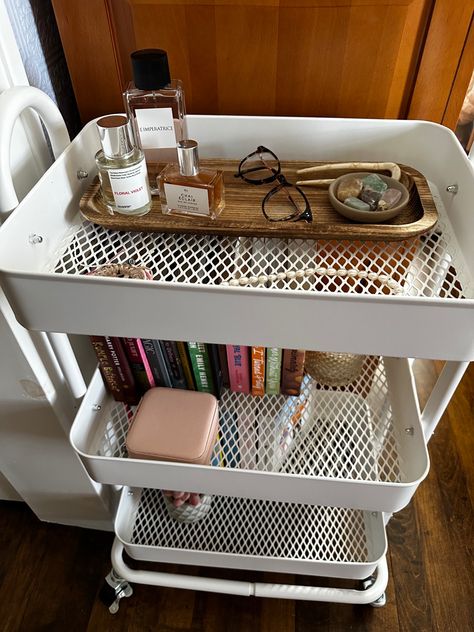 White book cart, books, perfume, crystals White Cart Decor, Nightstand Cart, Ikea Cart Ideas Bedroom, Cart Organization Ideas Bedroom, Cart As Nightstand, Storage Cart Aesthetic, Raskog Cart Bedroom, Trolley Cart Ideas Bedroom Aesthetic, Book Cart Decor Ideas