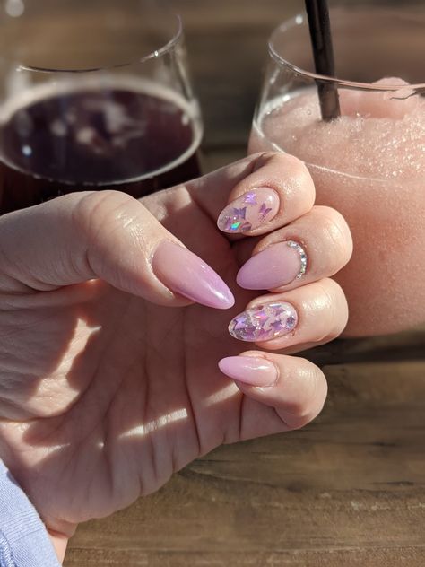 Almond shaped acrylic nails, mid length: Nude pink to pink lilac ombre nails on thumb, middle finger, and pinky finger. Middle finger has crest of tiny iridescent gemstones at the base. Pointee finger is nude pink to clear ombre with purple iridescent butterfly confetti and glitter. Ring finger is clear acrylic with same butterflies and glitter as pointer finger. Background is a wooden table with two stemless spill resistant wine glasses one with a dark beer, the other with a frozen rosé. Butterfly Inspired Nails, Lilac Nails With Butterflies, Lilac Butterfly Nails, Speak Now Nails, Lilla Nails, Butterflies Nails Acrylics, Gemstone Nails, Nails With Butterflies, Nails Extension