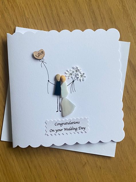 "Handmade Wedding Day Card  Decorated with coloured glass, paper flowers and wooden heart Give the marrying couple a beautiful and unique pebble card to treasure forever Measures 5 x 5\" (12.7 x 12.7cm) and comes with a white envelope Blank inside so you can write your own personal message Message text on tag can be changed to a more personalised one of your choice This card is made to order and although I always try to make others as close as possible to the one pictured here, this is not always possible due to the variation of the pebbles in size, shape and colour." Simple Wedding Cards Handmade, Handmade Wedding Cards Ideas Beautiful, Wedding Day Cards Handmade, Wedding Cards Handmade Congratulations, Diy Wedding Cards Handmade, Handmade Wedding Cards Ideas, Unique Wedding Card Design, Pebble Cards, Handmade Wedding Cards