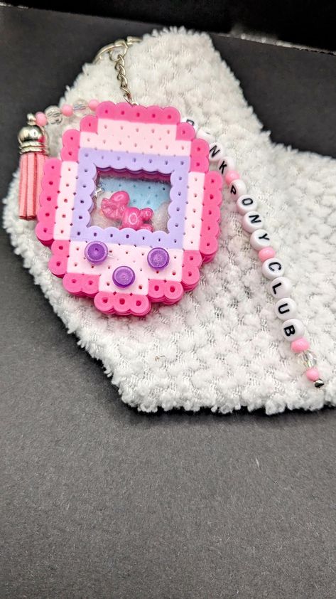 Add a nostalgic touch to your accessories with this handmade Tamagotchi-inspired keychain, crafted from vibrant Perler beads. Featuring a pink and purple color scheme with intricate details, this keychain also includes a delicate beaded tassel and "Pink Pony Club" charm for added flair. Perfect for fans of retro gaming or anyone looking to add a cute, whimsical piece to their collection. Clip it onto your keys, backpack, or purse for a playful pop of color wherever you go! Perler Bead Shaker, Pixel Art Keychain, Perler Bead Cute, Perler Beads Keychain, Perler Bead Magnets, Perler Bead Keychain, Perler Bead Designs, Pink Pony Club, Perler Creations