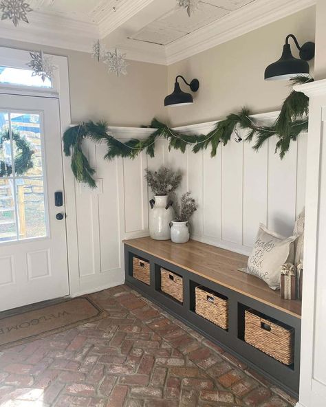 This entryway features cream-colored walls, white wainscoting, and a red brick herringbone floor. Branches in white jugs are placed on a long entryway bench with storage. Two black sconces illuminate the garland draped over the white wainscoting. Entryway Ideas Long Entry Hall, Brick Herringbone Floor, Entryway Wainscoting Ideas, Wainscoting Ideas Entryway, Long Entryway Bench, Entryway Shiplap, Brick Entryway, Black Entryway Bench, Wainscoting Entryway