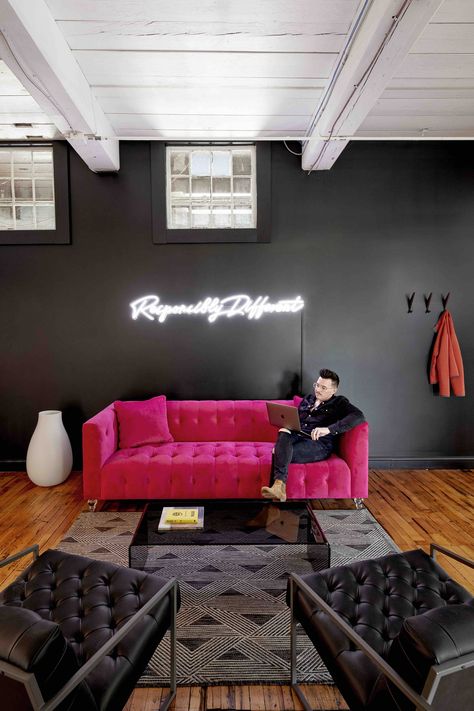 This moody office space pairs slate gray walls with a hot pink tufted couch, black leather chairs, and a black plexiglass coffee table for fun, retro vibes. A neon sign glows brightly, boasting the company's motto. Original glazed windows let in a modest amount of light, however, exposed ceiling beams keep the space feeling light and airy. Original stained hardwood floors give the room a homey feel. Black And Pink Salon, Maine Interiors, Hot Pink Couch, Moody Office Space, Plexiglass Coffee Table, Black Office Decor, Painted Ceiling Beams, Moody Office, Couch Black