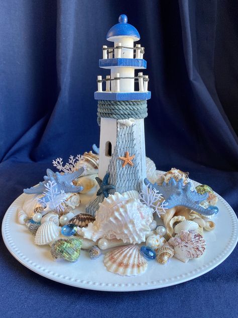 Tall Wood Lighthouse & Shells Nautical Centerpiece w/Slate Blue Starfish, Sand Dollar, Driftwood, Pearls, on White Charger The lighthouses have different designs on the netting, chosen at random. This one of a kind centerpiece is a special vignette to grace your coffee table, dining table, bringing a floral beach feeling to your decor. Or it would be so nice on the entry table to announce to your guests that beach lovers live here. It would make a charming addition to your patio table as well, but please bring back inside after use as it is not weather resistant. The shells in this arrangement are permanently set in place. Please remember that sea shells are delicate and fragile and can also have sharp edges. Use caution with children. While this creation may be used outside, it is not mea Seashell Table Decor Centerpieces, Nautical Centerpiece Ideas, Nautical Centerpieces, Beach Arrangements, Beach Theme Centerpieces, Nautical Centerpiece, Wood Lighthouse, Shell Centerpieces, Driftwood Centerpiece