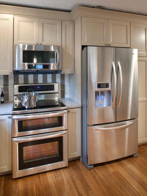 Neutral kitchen cabinets provide convenient storage and a clean facade in this kitchen. Stainless steel appliances, including double ovens, add a hint of shine to the muted color palette. Black and white subway tile add visual interest behind the stove with alternating vertical stripes. Fridge Next To Stove, Affordable Kitchen Appliances, Neutral Kitchen Cabinets, Fridge Cabinet, Hardwood Floors In Kitchen, Kitchen Appliance Storage, Refrigerator Cabinet, Kitchen Appliance Packages, Neutral Kitchen