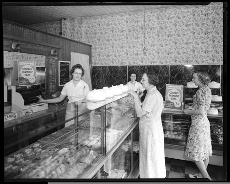 Horizontal, black and white photograph of an interior view of a bakery. Two employees, both women, stand behind the display counter, upon which is a line of cakes and also displays various pastries and other baked goods. Two customers, an older woman and a younger, are looking at the baked goods on display. A sign on the counter advertises the Coconut Harmony Cakes. 1940s Bakery, Danish Bakery, Donut Shops, Commercial Photography Studio, Vintage Bakery, Bakery Interior, Display Counter, Interior View, Black And White Photograph