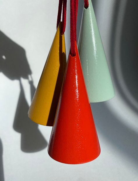 A set of three wooden, cone shaped hand painted Christmas tree ornaments in colors of mustard yellow, bright red, and eggshell blue are pictured with a white background. These three Christmas ornaments are held up by vegan red leather cords strung through the tops of each ornament. A shadow of the hand holding the ornaments is seen in the background. Bright Christmas Ornaments, Colorful Minimalism, Modern Christmas Ornaments, Eggshell Blue, Cone Trees, Modern Christmas Tree, Bright Christmas, Wood Christmas Ornaments, Wood Christmas