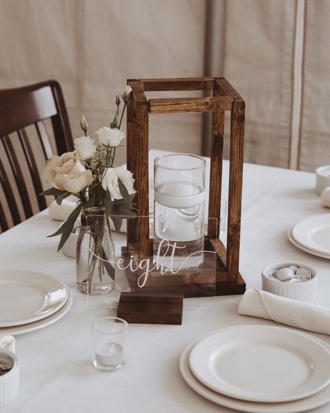 wedding guest table decor ideas rustic simple DIY hand made wooden lanterns with floating candle in vase milk jar with bud flowers centerpiece acrylic see through table numbers with wood base simple center piece decor ideas Floating Candle Lanterns, Floating Candles On Wood Centerpieces, Rustic Elegant Wedding Table Settings, Wood Lantern Table Centerpieces, Rustic Floating Candle Centerpieces, Wood Frame Centerpiece Wedding, Wood Lanterns Wedding Centerpiece Ideas, Wooden Lantern Wedding Centerpieces, Centerpiece Base Ideas