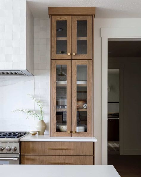 Warm wood kitchen cabinets and drawers contrast with the white walls in this home. A white tile backsplash and white countertops run the length of the wall, as well as a large square hood vent tiled in the same material as the backsplash. The glass cabinet doors feature brass knobs. Square Hood Vent, Warm Wood Kitchen Cabinets, Warm Wood Kitchen, Light Wood Kitchen Cabinets, Kitchen Cabinets And Drawers, Glass Kitchen Cabinet Doors, Light Wood Kitchens, White Tile Backsplash, Hood Vent