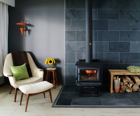 Black woodstove with large dark gray slate hearth, contrasting light tile flooring. Gray walls. Firewood box on side with pillow on top. Sophisticated design with just a touch of the rustic in the wood box. I love the orange lamp that echoes the fire and the orange in the kindling holder. Very pleasing. Mudroom With Wood Stove, Wood Stove Tile Wall, Slate Woodstove Hearth, Tile Behind Woodstove, Tile Under Wood Stove, Basement Wood Stove, Corner Wood Stove, Wood Stove Surround, Hearth Pad