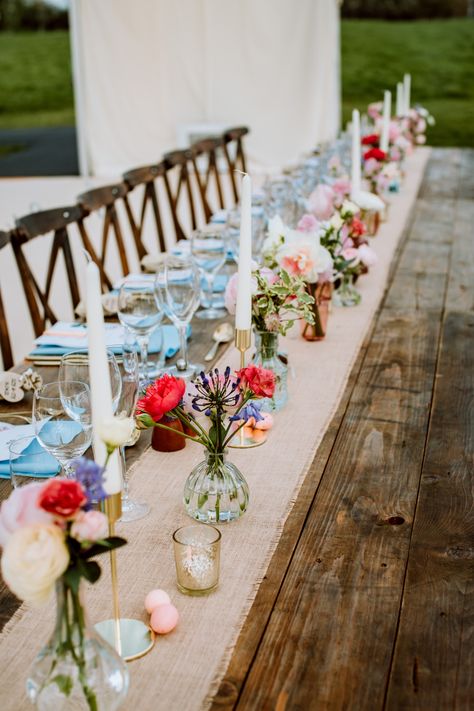 Pops of colour in low bud vases worked really well with the hessian runner and natural wooden tables for this Easter wedding reception in a clear span marquee at Barnsley House in the Cotswolds, UK. We add tealight votives in mercury gold glass, slim dinner candles and miniature chocolate eggs to complete the styling. Spring Wedding Long Table Decor, Wedding Tables With Bud Vases, Long Table Centrepiece Wedding, Long Wooden Wedding Table Decorations, Flowers Candles Centerpiece, Mix And Match Wedding Tables, Rustic Wedding Table Flowers, Wedding Table Decorations Wooden Table, Wedding Table Tealights