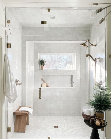 A wood bench is placed behind the glass panels of a shower with a window. The window is surrounded by white marble tile and a rectangular shower niche. Nickel shower heads are anchored to one wall. Subtle nature decor accessorizes a modern black table. Honey Marble Bathroom, Zia Tile Bathroom, Bathroom Gold Fixtures, Vanilla Bathroom, Dr Bathroom, Gold And White Bathroom, Shower Windows Ideas, White And Gold Bathroom Decor, White And Gold Bathroom