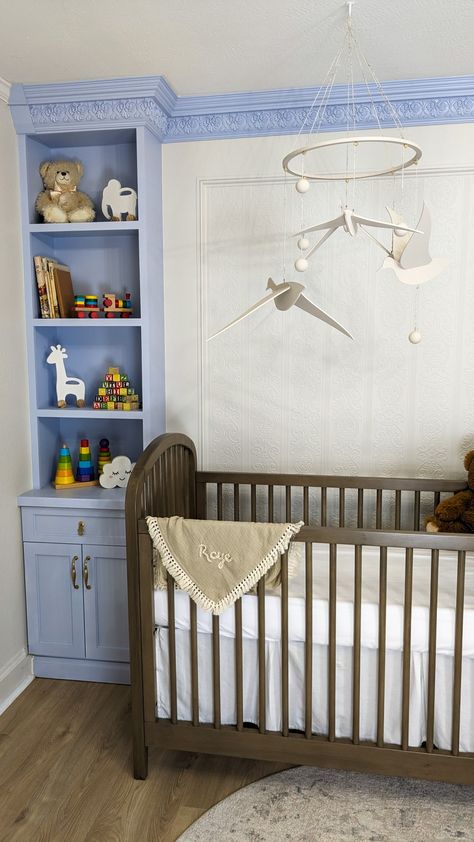 The left corner of a nursery. In the corner there are periwinkle floor to ceiling built in shelves. The crown molding connects with the shelves and is also periwinkle. The walls are covered in textured wallpaper. In front of the wall is a brown crib with a neutral rug beneath it. Hanging over the crib is an off-white mobile with birds and spheres on it. Built In Crib, Molding Wallpaper, Nursery Room Diy, Shelves Nursery, Nursery Room Inspiration, Diy Nursery, Built In Shelves, Crown Molding, Baby Room